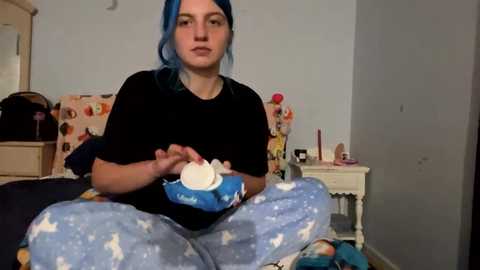 Media: A video shows a young person with blue hair, wearing pajamas, seated on a bed in a messy bedroom. They hold a blue cupcake container.