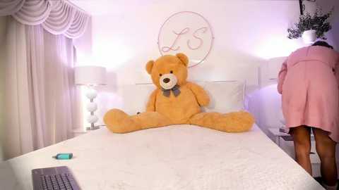 Media: Video of a plush teddy bear sitting on a white bed in a pastel-themed bedroom with a pink skirted woman standing nearby, a laptop on the bed, and a white lampshade.