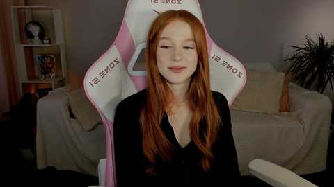 Media: Video of a fair-skinned, long-haired redhead in a pink gaming chair, wearing a black top, in a dimly lit living room with beige furniture, shelves, and a plant.