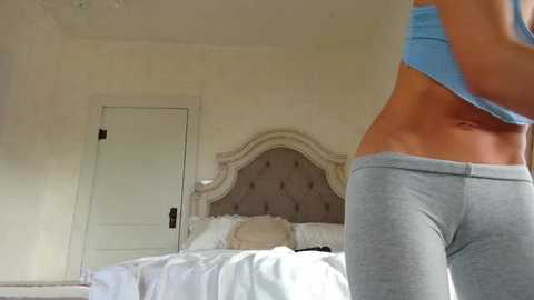 Media: Video of a woman in a light blue sports bra and gray leggings, lifting her shirt to reveal toned abs, standing beside a neatly made bed with white linens and a tufted headboard.