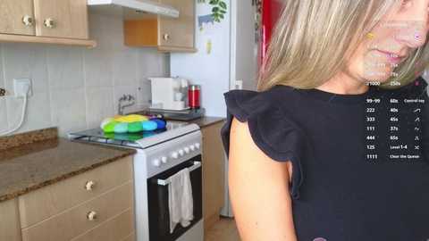 Media: Video of a blonde woman in a black sleeveless top, standing in a modern kitchen with light wood cabinets, granite countertops, and a colorful rainbow cake on the stove.