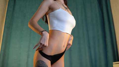 Media: Video of a slim, light-skinned woman with long brown hair wearing a white crop top and black panties, standing against teal curtains. She has a tattoo on her left thigh.