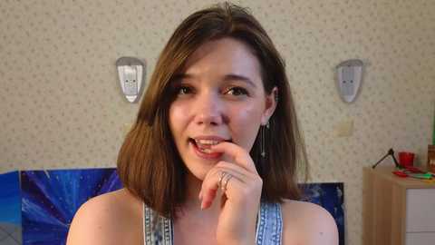 Media: Video of a young woman with shoulder-length brown hair, wearing a sleeveless blue top, smiling and biting her finger in a playful manner. Background features a wallpapered wall with a blue pattern, a wooden dresser, and a blue bedspread.