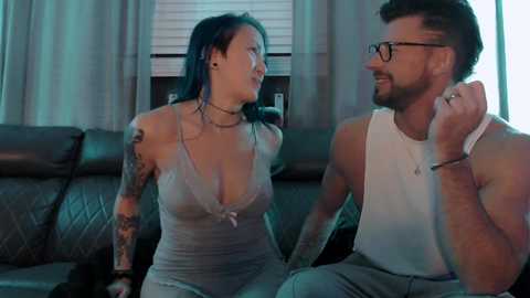 Media: A video of a tattooed woman in a gray dress and a man in a white tank top and glasses, sitting on a couch in a dimly lit living room.