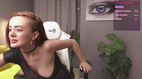 Media: Video of a young woman with pale skin and red hair, wearing a black choker and top, leaning back on a white Artix chair. Background shows a green plant, gray wall, and a monitor displaying Twitch chat.