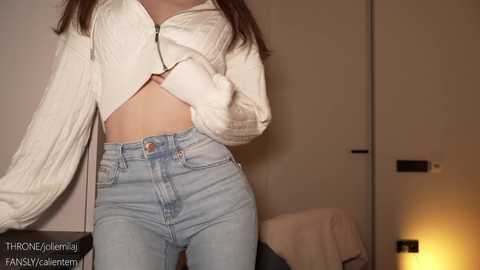 Media: Video of a slender, light-skinned woman with long dark hair, wearing a white cropped sweater and high-waisted light blue jeans, standing indoors against a neutral-toned wall.