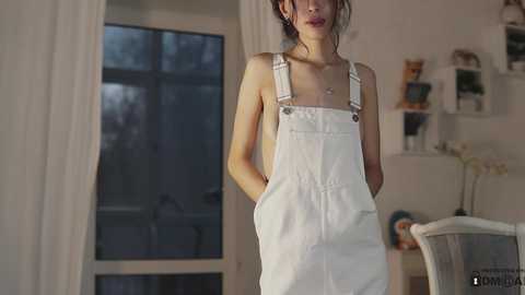 Media: Video of a slim, light-skinned woman with shoulder-length hair wearing white overalls, standing in a softly lit room with a large window and bookshelves.