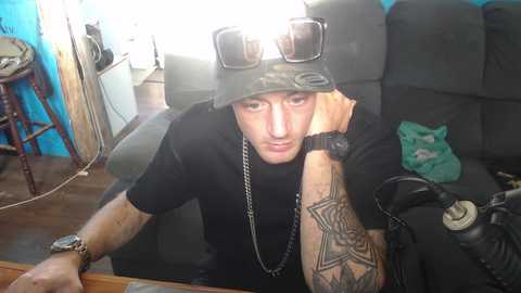 Media: A video of a tattooed man in a black T-shirt, wearing aviator sunglasses and a camo bucket hat, leaning on a table with a tattooed arm. Background shows a blue wall, a green bag, and a chair.