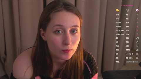 Video of a pale-skinned, young woman with long, straight brown hair, wearing a black and pink striped sweater, sitting indoors with beige curtains and a computer screen displaying a chat interface in the background.