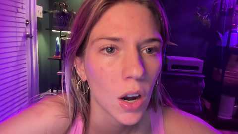 Media: Video of a young Caucasian woman with light skin, wet blonde hair, and a surprised expression, wearing a pink tank top, set in a dimly lit room with purple lighting, shelves, and a window in the background.