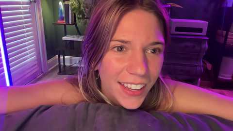 Media: Video of a smiling Caucasian woman with light brown hair, wearing a gray top, lying on a bed in a dimly lit room with purple and green lighting.