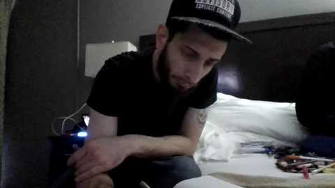 Media: Video of a bearded man with headphones on, wearing a black cap and t-shirt, sitting on a bed in a dimly lit room with disheveled bedding.