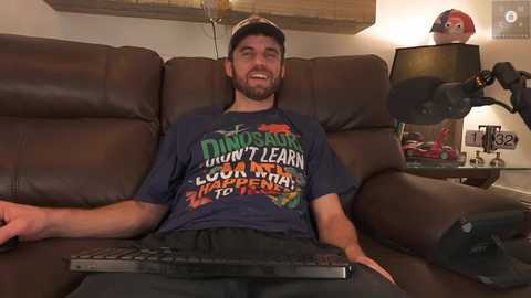 Media: Video of a bearded man in a blue T-shirt with dinosaur-themed text, sitting on a brown leather couch, holding a remote.