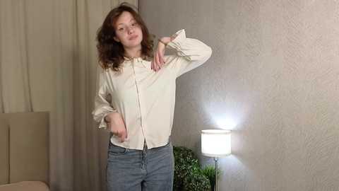 Media: Video of a white woman with curly hair, wearing a cream blouse and blue jeans, adjusting her blouse, standing in a beige, textured-walled room with a lamp and potted plant.