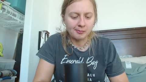 Media: Video of a young Caucasian woman with light skin and shoulder-length blonde hair, wearing a dark gray t-shirt with white text, sitting in a bedroom.