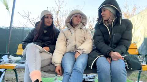 Media: Video of three young women sitting on a bench outdoors, bundled up in winter coats and hats, with bare trees and a clear sky in the background.