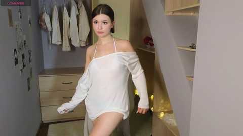 Media: Video of a young woman with pale skin and dark hair, wearing a loose, white, off-shoulder sweater, standing in a cluttered room with clothes hanging, a wooden dresser, and a spiral staircase.