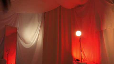 Media: Video of a dimly lit room with white drapes, a red-lit lamp, and a reflective surface, creating a warm, mysterious ambiance.