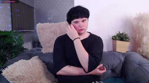 Media: Video of a young man with pale skin, dark hair, and a black shirt, sitting on a grey couch with beige pillows, holding his head, in a cozy, dimly lit room with plants and wall art.