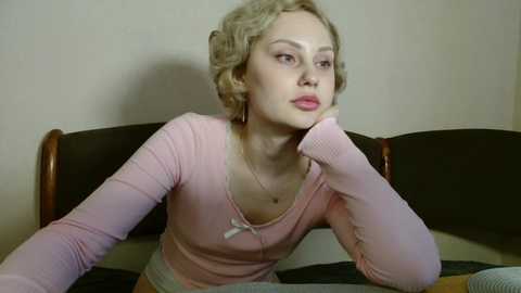 Media: Video of a blonde woman with wavy hair, wearing a pink long-sleeve top, leaning on a sofa, looking contemplative.