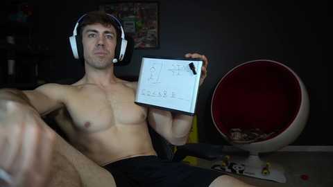 Media: Video of a shirtless, muscular young man with short brown hair, wearing headphones, holding a whiteboard with sketches, in a dimly lit room with a red egg chair and scattered toys.