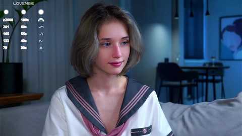 Media: Video of a young, light-skinned woman with shoulder-length, wavy blonde hair in a white sailor-style top, sitting indoors with a serene expression, surrounded by a modern, dimly-lit living room with minimalist decor.