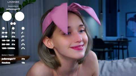 Media: Video of a young woman with fair skin, shoulder-length blonde hair, and pink bunny ears, smiling in a dimly-lit room with a white couch and a potted plant.