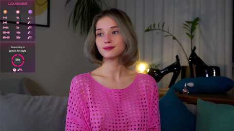 Media: Video of a young Caucasian woman with short blonde hair, wearing a pink crochet sweater, standing in a cozy, dimly lit living room with a calendar, potted plants, and a lamp.