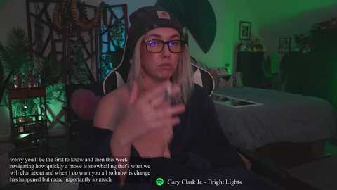 Media: A video shows a young, light-skinned woman with long, platinum blonde hair and glasses, wearing a black hat and jacket, sitting on a gaming chair in a dimly lit room.