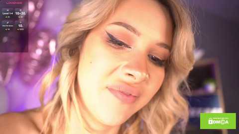 Media: Video of a smiling, fair-skinned woman with wavy blonde hair, wearing subtle makeup, in a softly lit, blurred indoor setting.