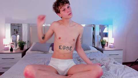 Media: A video of a young, slender, shirtless man with short brown hair, tattoos on his chest, wearing white briefs, sitting cross-legged on a bed in a modern bedroom with mirrored headboard and bedside tables.