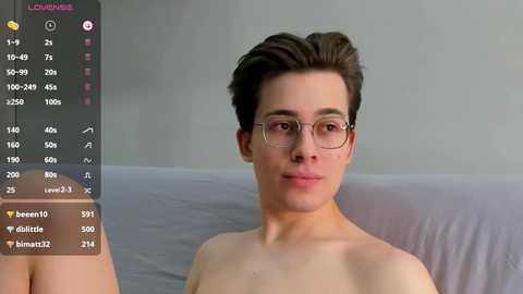 Media: Video of a young, fair-skinned man with short brown hair, wearing glasses, sitting shirtless on a gray couch, with a digital screen overlay showing his stats.