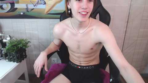 Media: Video of a young, shirtless, skinny, fair-skinned male with short hair, wearing black Calvin Klein underwear, seated on a black gaming chair in a modern, beige-tiled room with a green plant and colorful abstract art in the background.