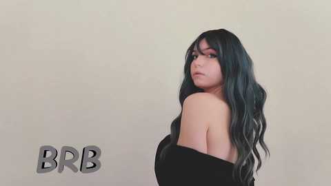 Media: Video of a young woman with long, wavy, dark green hair, wearing an off-shoulder black dress, against a plain beige background. \"BBR\" text is in the lower left corner.