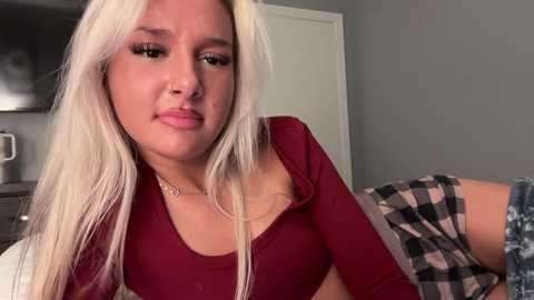 Media: Video of a young woman with long, platinum blonde hair, wearing a maroon long-sleeve top, sitting on a bed in a dimly lit room with a gray wall and a checkered blanket.