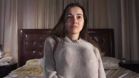 Media: Video of a young woman with long brown hair, wearing a cream-colored ribbed sweater, standing in a dimly-lit bedroom with a dark leather headboard, beige curtains, and a bed with a striped blanket.