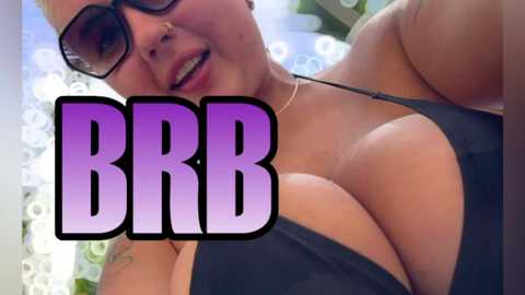 Media: Video of a busty, tanned woman with black-rimmed glasses, wearing a low-cut black bikini, smiling, with \"BBB\" text overlay. Background blurred, sunlit.