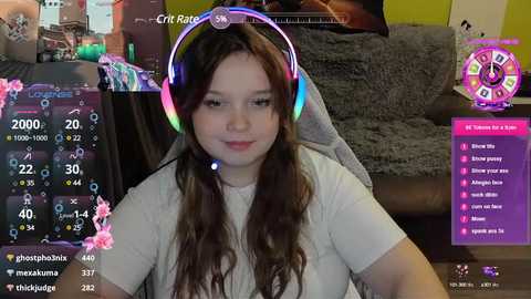 Media: A video of a young woman with long brown hair, wearing a white t-shirt, playing a video game. She has a headset on, surrounded by gaming interface elements.