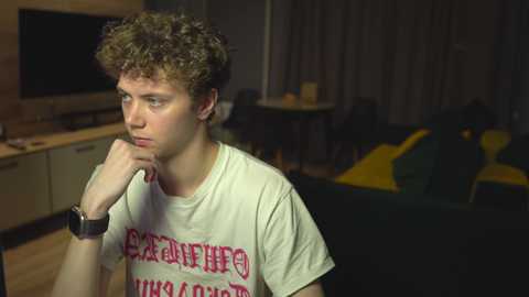 Media: Video of a young man with curly brown hair, wearing a white T-shirt with red text, sitting pensively on a dark couch. Background shows a dimly lit living room with a TV, a wooden shelf, and yellow pillows.