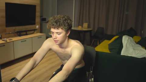 Media: Video of a shirtless, curly-haired young man riding an exercise bike in a modern, dimly lit bedroom. The room features a dark green bed with yellow pillows, a TV on a wooden cabinet, and beige curtains.