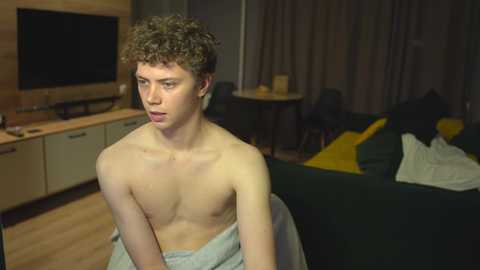 Media: Video of a shirtless, curly-haired, fair-skinned teenage boy sitting on a chair in a dimly lit, modern living room with a TV, wooden furniture, and yellow cushions.