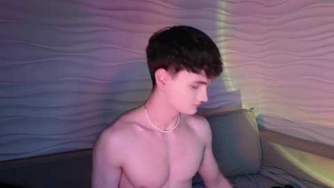 Media: Video of a young, shirtless, fair-skinned, slim Caucasian male with short dark hair, wearing a thin silver chain necklace, sitting on a bed with a textured, wavy wall background and soft, ambient lighting.