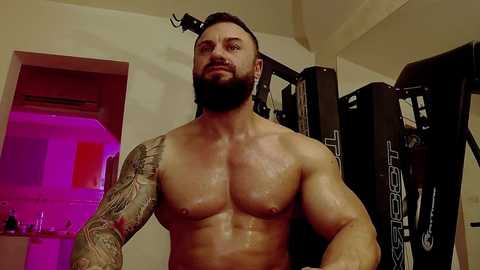 Media: Video of a muscular, shirtless man with a beard and intricate tattoos, standing in a gym. He has a light skin tone and a serious expression. Background includes gym equipment and a partially visible doorway with pink lighting.