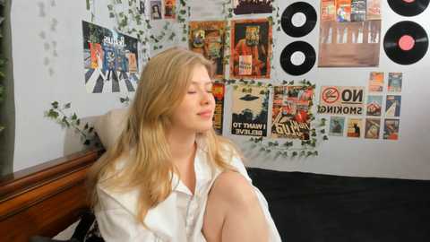 Media: Video of a blonde, fair-skinned woman with long hair, wearing a white shirt, sitting on a bed, surrounded by posters and vinyl records on a white wall.