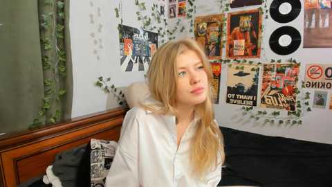 Media: Video of a blonde woman in a white shirt, sitting on a bed, with a wall covered in posters and records, and green vines.