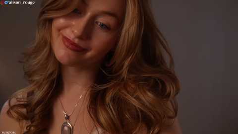 Media: Video of a smiling Caucasian woman with wavy, light brown hair and fair skin, wearing a gold necklace, against a blurred, dark background.