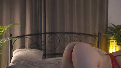 Media: Video of a woman in red lingerie on a bed with a wrought iron headboard. Her back is arched, revealing her bare buttocks. The background features gray curtains and green potted plants.