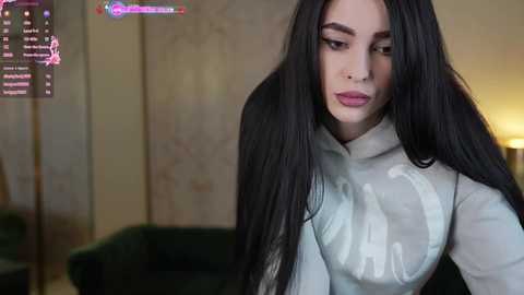 Media: Video of a woman with long black hair, fair skin, wearing a light grey sweater with a white logo, standing indoors in a dimly lit room.