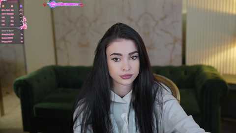 Media: A video of a young woman with long black hair, wearing a light grey hoodie, seated in a dimly lit room with a green velvet sofa and vertical blinds in the background.