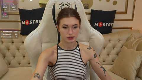 Media: Video of a fair-skinned woman with auburn hair, tattoos, wearing a black and white striped halter top, seated in a white gaming chair with \"MST3K\" logo, against a beige tufted sofa with pillows, in a cozy, well-lit room.